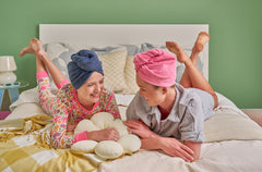 Two young girls laying on a bed, each wearing a microfiber Turbie twist. The girl on the left is wearing a dark blue Turbie twist hair towel, and the girl on the right is wearing a pink Turbie twist.